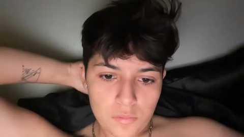 A video of a young, fair-skinned man with short, dark hair and a neutral expression. He's shirtless, wearing a gold chain necklace, and has a tattoo on his left arm. He's lying on a black pillow.