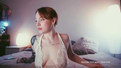 Video of a fair-skinned woman with light brown hair in a bun, wearing a revealing white lace halter top, sitting on a bed in a dimly lit bedroom.