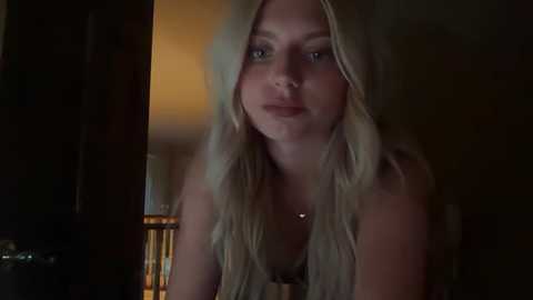 Video of a young woman with long, straight, platinum blonde hair, fair skin, and light blue eyes, wearing a black bra. She stands indoors, dimly lit, with a wooden door partially open behind her.