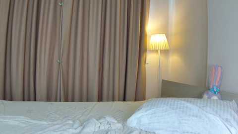 Video of a minimalist bedroom with beige curtains, a white bed with a textured blanket, a nightstand holding a lamp, and a colorful stuffed animal.