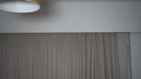 Video of a beige, pleated curtain hanging from a white ceiling track. The ceiling is painted white, and a yellow lampshade is partially visible in the top left corner.
