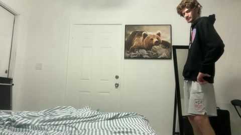 Video of a young man with curly brown hair in a black hoodie and grey shorts, standing beside a bed with a white door and a framed bear painting in a minimalist bedroom.