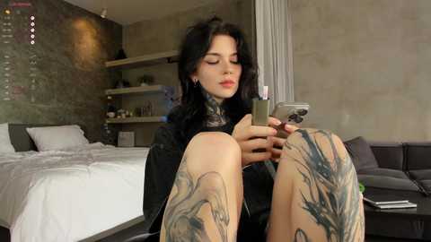 Video of a woman with dark hair, tattoos, and black clothes sitting on a bed, looking at a phone, in a modern, minimalistic bedroom.