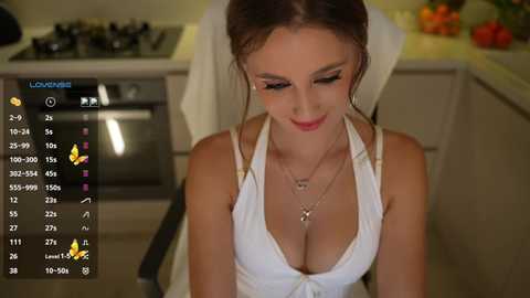 Video of a young woman with light skin, brown hair, and large breasts, wearing a revealing white halter top, seated in a modern kitchen with colorful fruits and a stove in the background.