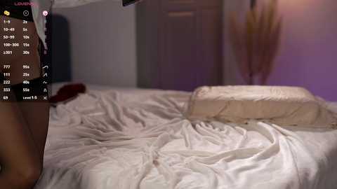 Video of a dimly lit bedroom with rumpled white sheets, a beige pillow, and a blurred figure partially visible in the background.