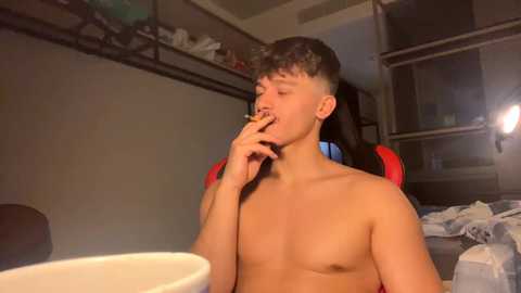 Video of a shirtless young man with short brown hair, smoking a cigarette, seated at a cluttered desk in a dimly-lit room with a bed and shelves in the background.
