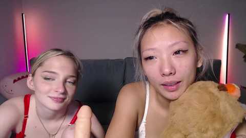 Video of two young women, one blonde with fair skin and the other with darker hair, both smiling, holding teddy bears, sitting on a gray couch with pink and purple lighting in the background.