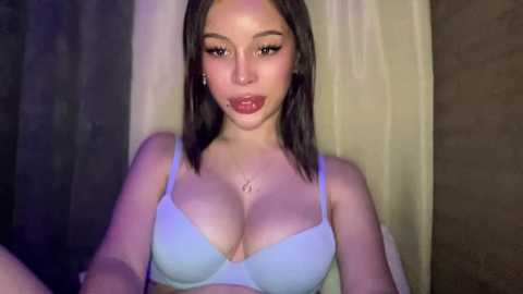 Video of a young woman with fair skin, straight brown hair, wearing a light blue bra, sitting in a dimly lit room with beige curtains.