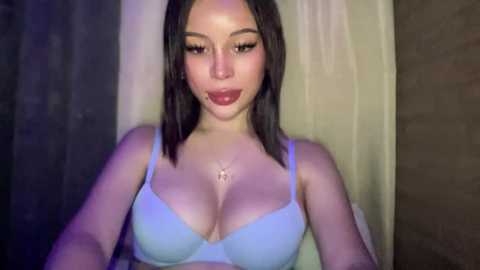 Video of a young Asian woman with straight dark hair, wearing a light blue bra, taking a selfie in a dimly lit room.