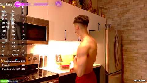 Video of a shirtless, muscular young man in red shorts cooking in a modern kitchen with stainless steel appliances, beige tiled walls, and a white microwave.