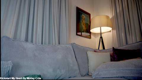 A cozy living room with a plush, light blue sofa adorned with striped pillows, a white table lamp casting a warm glow, and framed artwork on the wall.