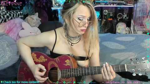 A young, fair-skinned, blonde woman with glasses, wearing a black bra and multiple chokers, strums a red electric guitar on a bed, surrounded by a messy room with blue bedding and various objects.
