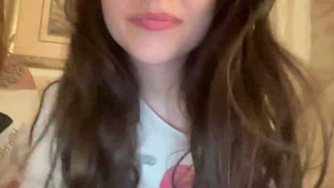 A close-up video of a young woman with long, wavy brown hair and light skin, wearing a white T-shirt with a red rose graphic. The background shows a blurred, beige wall with framed artwork.