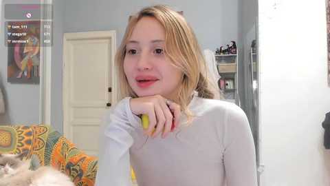 Video of a young blonde woman with fair skin, wearing a white long-sleeve top, sitting on a colorful patterned couch, looking at a camera. Background includes a closed white door, a mirror, and a cat.