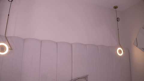 Video of a minimalist room with white walls and textured paneling. Two modern, circular pendant lights hang from the ceiling, casting a soft, warm glow. The room's decor is simple, with no visible furniture.