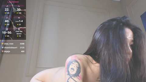 Video of a nude woman with long black hair, facing away, showing a colorful tattoo on her left shoulder. She stands in a dimly lit room with a digital camera viewfinder overlay showing stats.