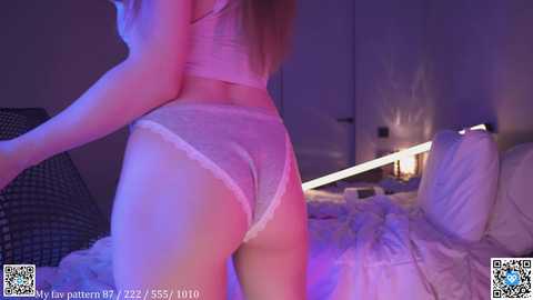 Video of a woman with long, blonde hair wearing sheer white lace lingerie, standing on a bed in a dimly lit, purple-lit bedroom.