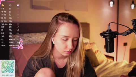A video of a blonde woman with a contemplative expression, wearing headphones, sitting in a dimly lit bedroom. A microphone is positioned in front of her.