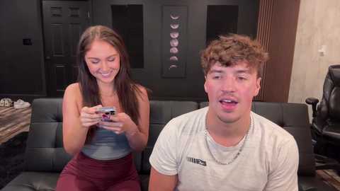Video of a young woman with long brown hair and a man with curly brown hair, both smiling while playing a video game on a black sofa in a modern, dimly lit room.
