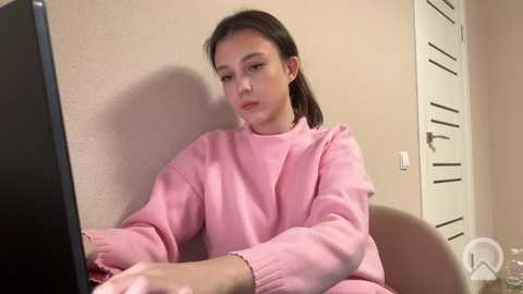 Video of a young woman with straight, dark hair, wearing a pink sweater, sitting at a computer desk in a beige room. She appears focused, with a neutral expression.