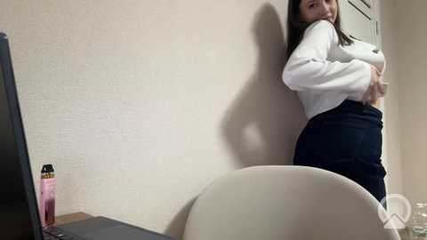 Video of an East Asian woman with long black hair, wearing a white sweater and dark pants, leaning against a beige wall, looking over her shoulder. A computer monitor and a water bottle are visible on a desk in the background.