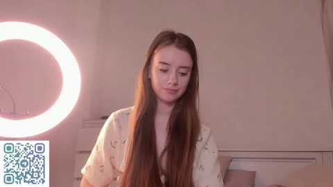 Video of a young Caucasian woman with long, straight brown hair, wearing a light-colored, patterned shirt. She stands in a softly lit room with a circular ring light to her left.