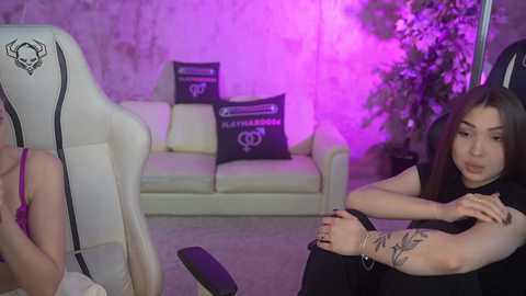 Video of a young woman with light skin and long dark hair, wearing a black sleeveless top and purple bracelet, sitting on a white gaming chair, with a tattooed arm. Background includes a white couch with purple pillows, a plant, and a purple-lit room.