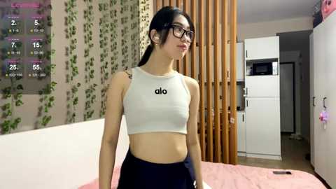 Video of a slim, young Asian woman with long black hair, wearing glasses and a white \"ado\" crop top, standing in a modern bedroom with wooden vertical panels, white cabinets, and a pink bedspread.