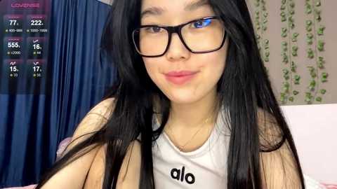 Video of an Asian woman with long black hair, glasses, and a white tank top, smiling in a cozy room with a blue curtain and green ivy wall.