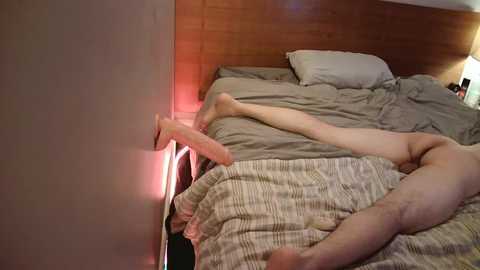 Video of a man, naked, lying on a bed with a beige and white striped comforter, his legs stretched out. The room has a wooden headboard and a bedside lamp, with red light from a lamp casting a soft glow.