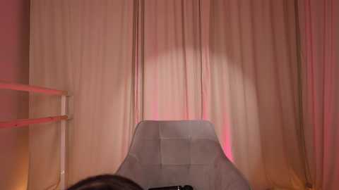 Video of a modern, minimalist room with a large, light gray, tufted leather chair centered against beige curtains. The room is dimly lit with soft pink and red lighting, creating a warm, cozy atmosphere.