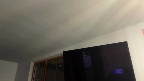 Video of a modern living room with a large, glossy black flat-screen TV mounted on a white wall. A wooden-framed window with reflective glass is visible to the left. The room has a minimalist, contemporary design with neutral color tones.