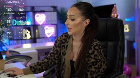 A video shows a woman with long brown hair, wearing a leopard-print jacket, seated at a desk, in a dimly-lit office. Virtual reality screens display numbers and a heart graphic.