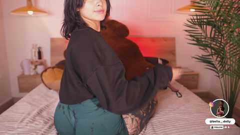 Video of a woman with short black hair, wearing a black sweater and teal skirt, sitting on a bed with a wooden headboard, holding a brown teddy bear, in a modern, warmly lit bedroom.