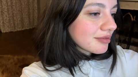 Media: A video of a young woman with pale skin, straight black hair, and a pink blush on her cheeks, wearing a white turtleneck sweater, seated indoors with a textured brown curtain in the background.