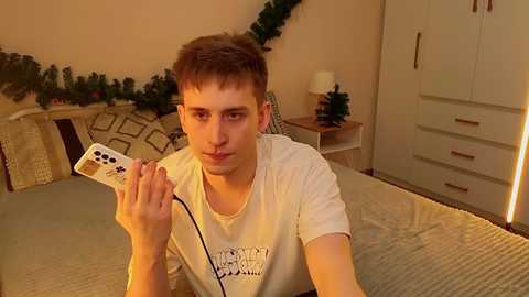 Video of a young Caucasian man with short brown hair, wearing a white T-shirt, holding a video game controller, in a cozy bedroom with a bed, dresser, and Christmas decorations.