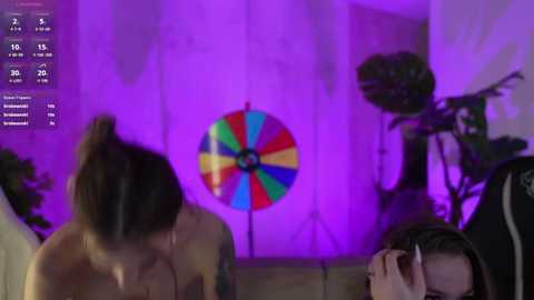 Video of a woman with dark hair tied up, facing a colorful pinwheel with purple lighting in a dimly lit room, another person's hand covering their face.