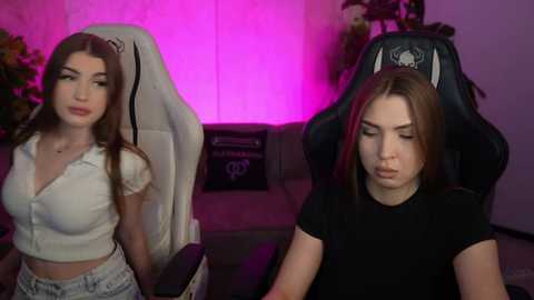 Video of two women, one in a white crop top, the other in a black t-shirt, sitting in gaming chairs with neon lights in the background, a plant on the left.