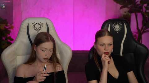 Video of two young women, one with long brown hair and another with red lipstick, sitting on gaming chairs in a dimly lit room with pink lighting and greenery.