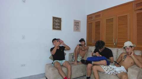 Video of four shirtless, young, fit men in casual clothes sitting on a beige sofa in a simple room with wooden shutters, drinking from wine glasses.