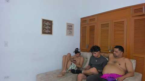 Video of three men in casual attire, one overweight, sitting on a beige couch in a cozy, minimalistic room with wooden shutters and coffee-themed decor.