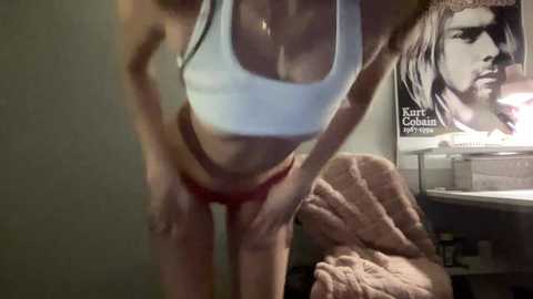 Video of a slender, fair-skinned woman with medium-sized breasts, wearing a white tank top and red panties, bending over, in a dimly lit room with a Nirvana poster and a beige blanket.