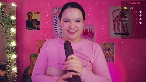 Video of a smiling Asian woman with dark hair, holding a large, black dildo. Pink room with posters, string lights, and a plush toy in the background.