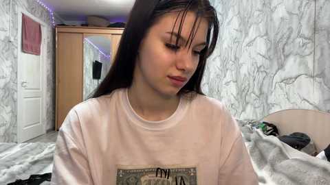 A video of a young woman with straight, dark hair, wearing a white T-shirt with a dollar bill graphic, sitting in a messy bedroom with white marble-patterned wallpaper, a wooden wardrobe, and a bed with scattered clothes.