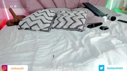 Video of a bed with white sheets, two black-and-white zigzag-patterned pillows, a remote control, and a hand holding a dildo. The background features a pink upholstered headboard and green lights.
