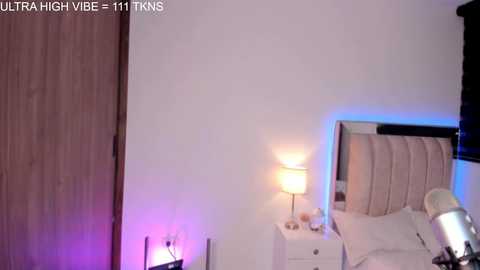 Video of a minimalist bedroom with a white bed, beige headboard, nightstands, and a lamp emitting blue and purple light.