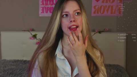 Media: Video of a young woman with long, straight blonde hair, wearing a white blouse, with a surprised expression and hand to her mouth, in a pink-themed bedroom.