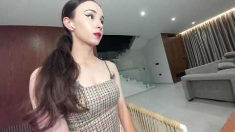 Video of a slender, fair-skinned woman with long brown hair, wearing a plaid dress, standing in a modern, minimalist living room with light wood and white decor.