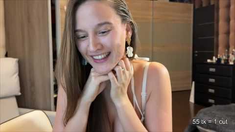 Video of a smiling, slender Asian woman with long, straight hair, wearing a white spaghetti-strap top, in a modern bedroom with wooden furniture and a bed.