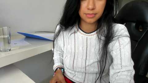 Video of a young woman with long black hair, wearing a white knit sweater, sitting at a desk with a glass of water, a blue folder, and a black chair in the background.